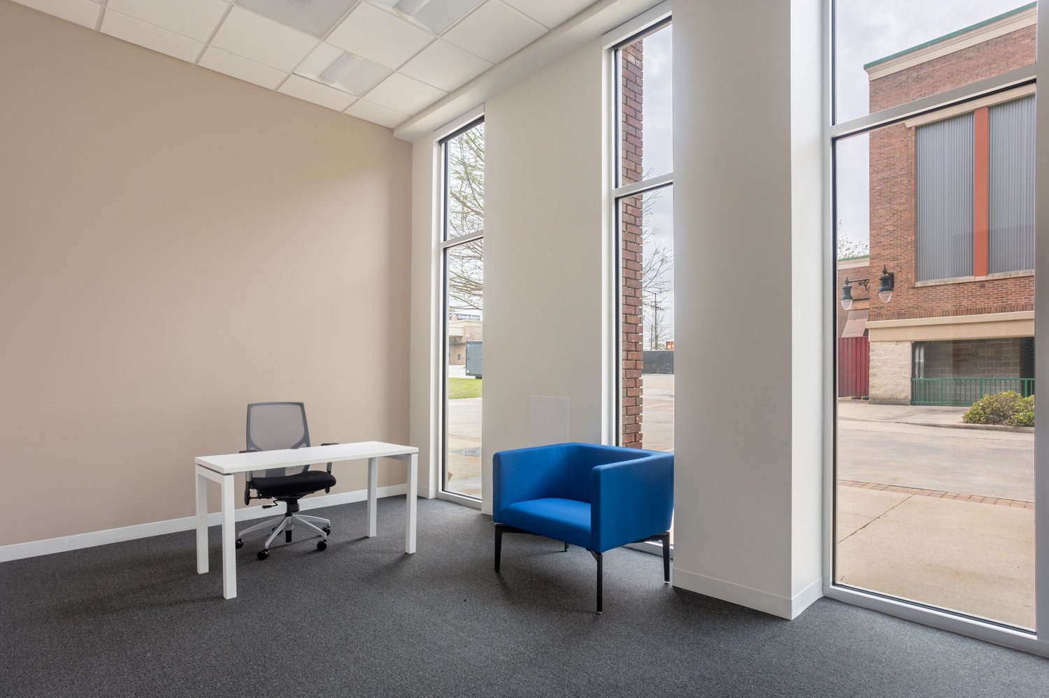 The image shows a modern office space with a white desk, a black office chair, and a blue armchair. The room has large windows that provide natural light and a view of the exterior. The walls are a neutral beige color, and the floor is covered with gray c