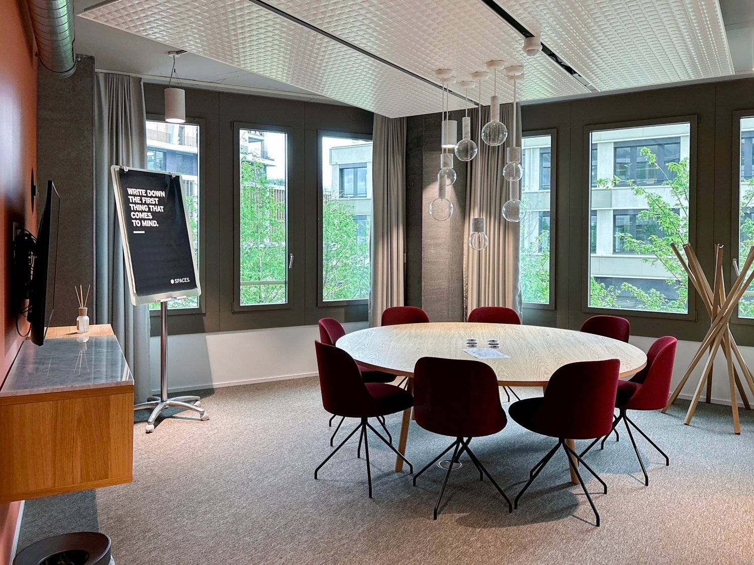 modern meeting room with round table, red chairs, large windows, wooden desk, blackboard, pendant lights, carpeted floor