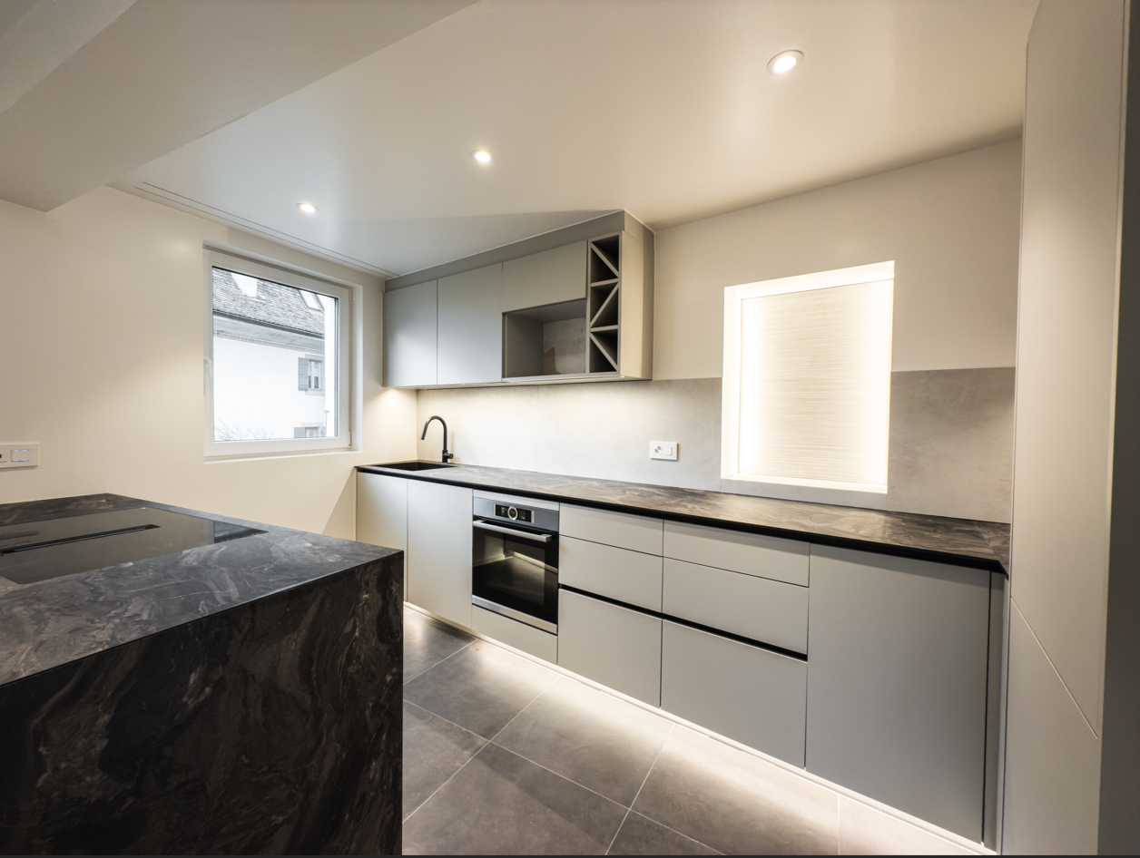 Spacious modern kitchen with gray cabinets, black marble countertops, built-in oven, and a dishwasher. The kitchen has recessed lighting and a large window providing natural light.