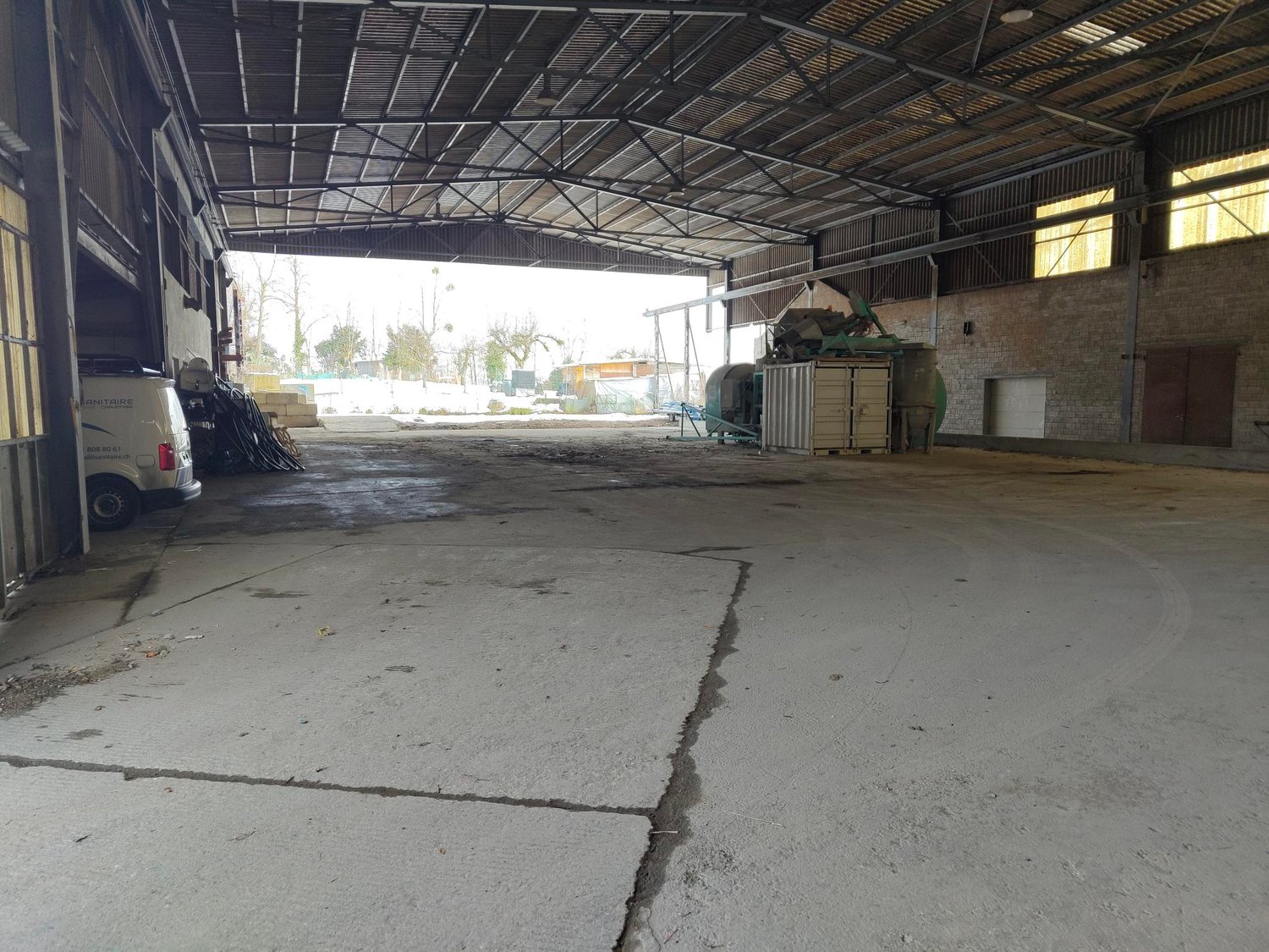 empty warehouse with concrete floor, metal roofing, large open space, some equipment, brick walls, windows, door on the right side