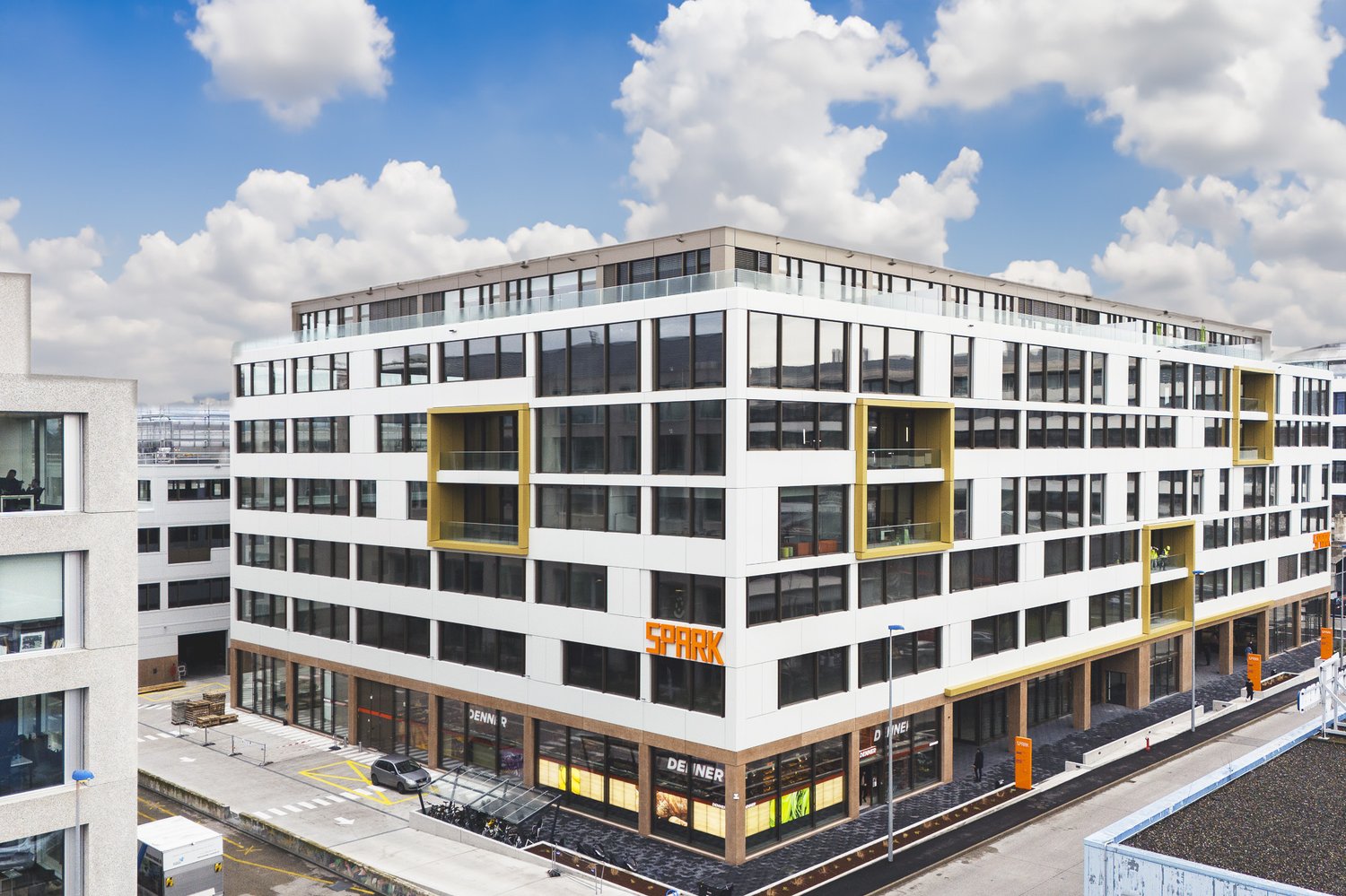 A modern, multi-story commercial building with a large glass facade and yellow accents. The building appears to have a parking area in front of it.