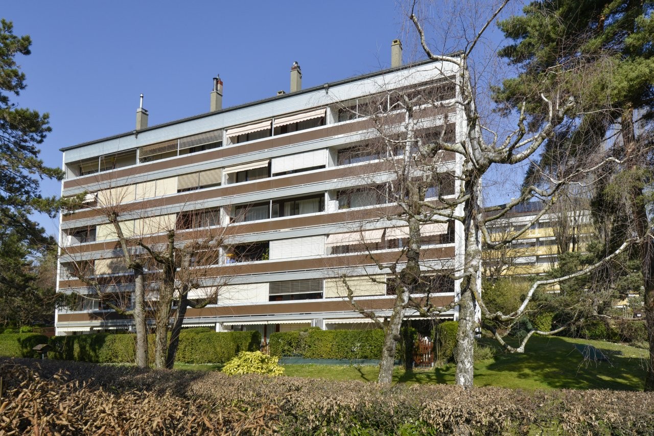 large building with many apartments, multiple floors, white exterior, brown accents, multiple windows, surrounded by greenery