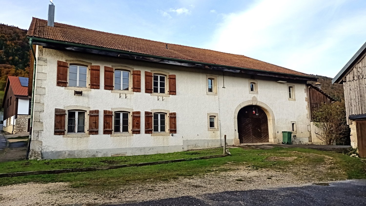 Two-story building with white exterior walls, red tile roof, and wooden shutters on the windows. The building has an arched entryway and is surrounded by a grassy area.