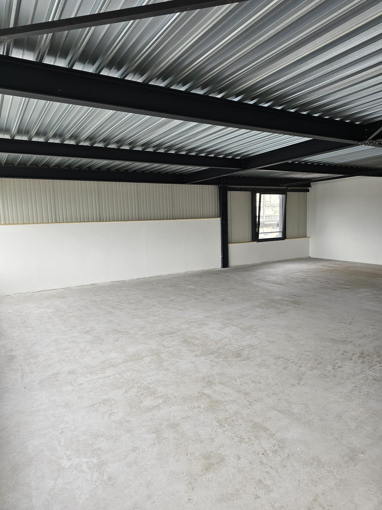 The image shows the interior of a warehouse or industrial space. The ceiling is made of corrugated metal panels, and there are black metal beams supporting the structure. The floor is a plain concrete surface. The space appears to be empty and unfinished,