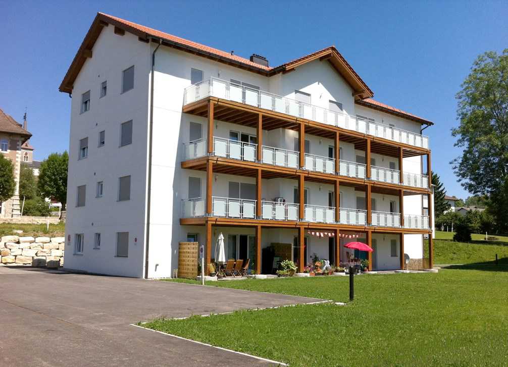 white apartment building, brown roof, balconies, outdoor tables, umbrellas, green lawn
