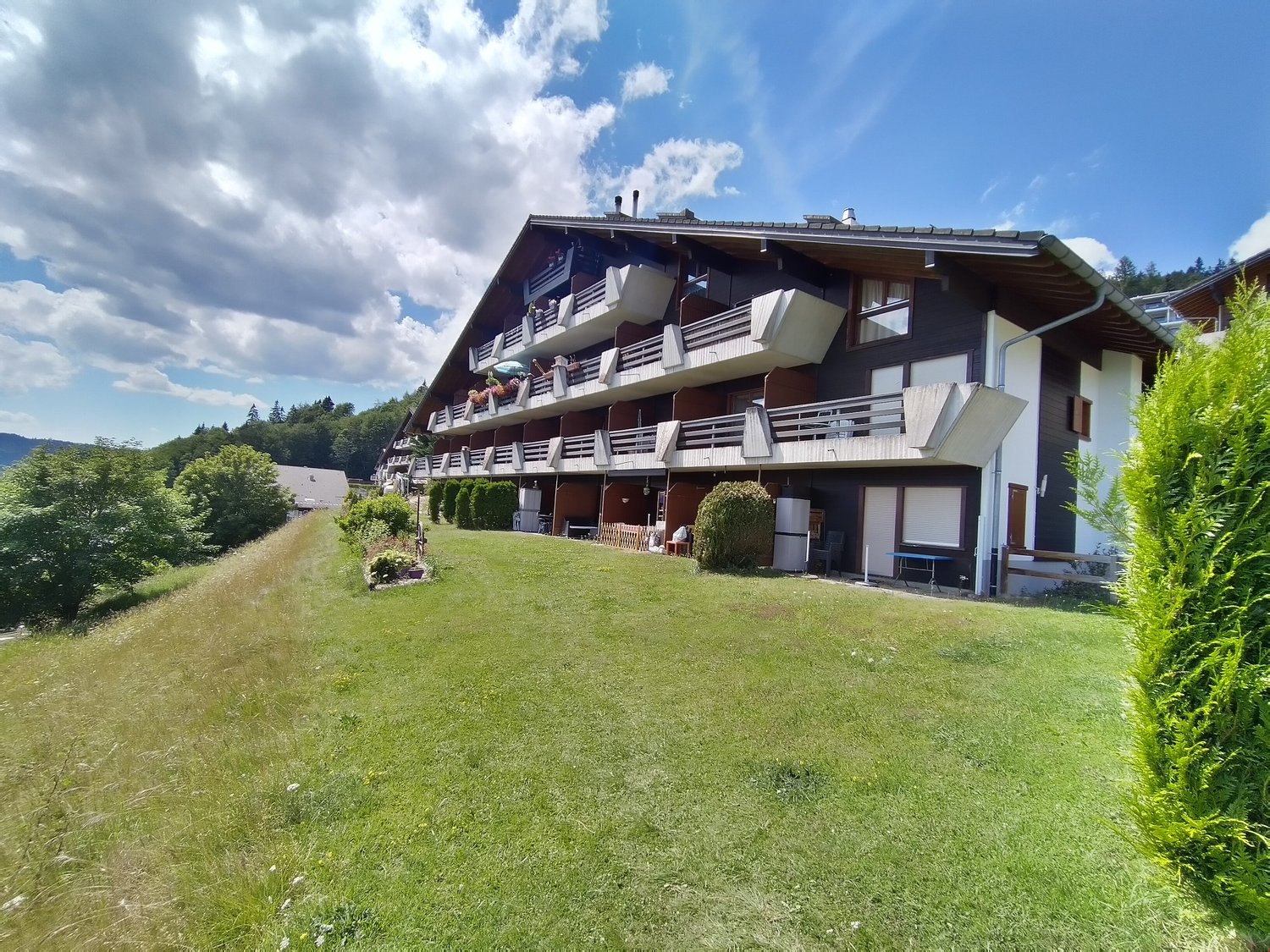 A multi-story chalet-style building with a sloped roof, wooden exterior, and balconies overlooking a grassy lawn surrounded by trees and mountains in the background.