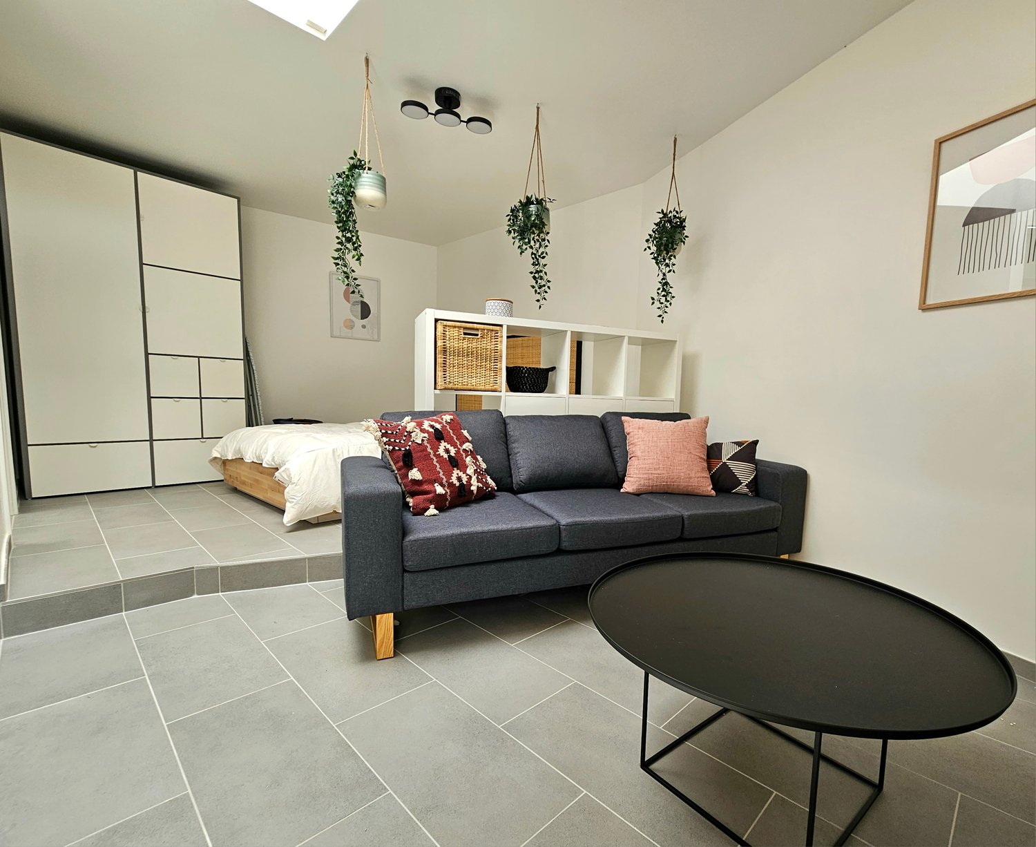 Open-concept studio apartment with a living area, sleeping area, and storage shelves. The room features a gray sofa, a black coffee table, and hanging plants. The floor is covered with gray tiles, and there is a sliding glass door leading to a balcony or