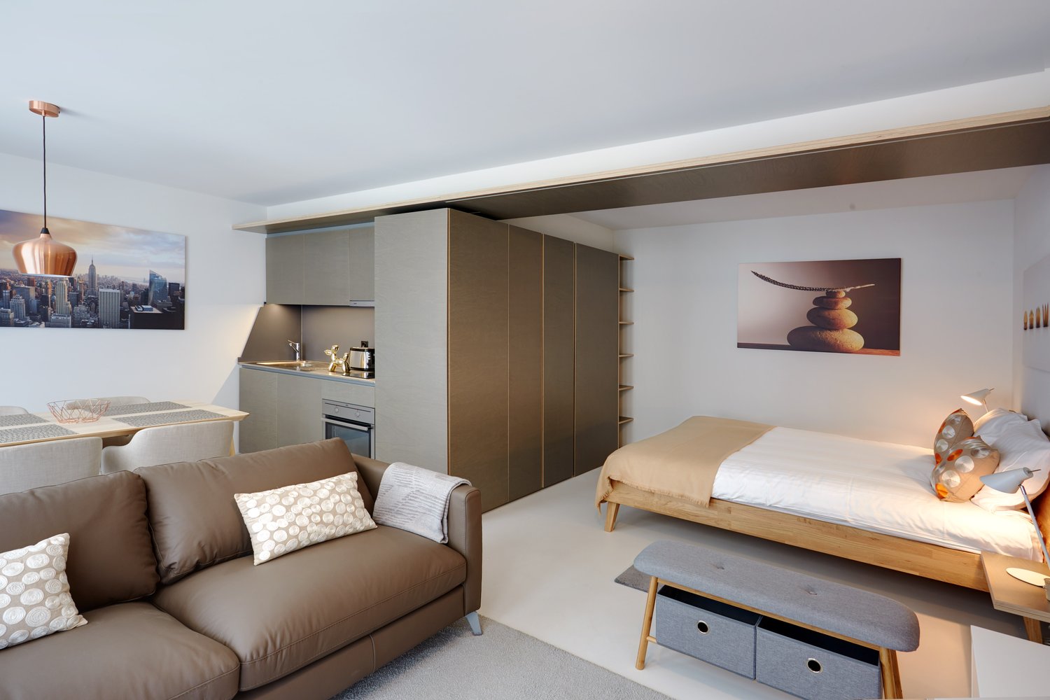 Open-plan studio apartment with living area, kitchenette, and sleeping area. Features include a sofa, coffee table, wall art, built-in storage, and a kitchenette with a dishwasher. The apartment has a modern, minimalist design with neutral colors and natu