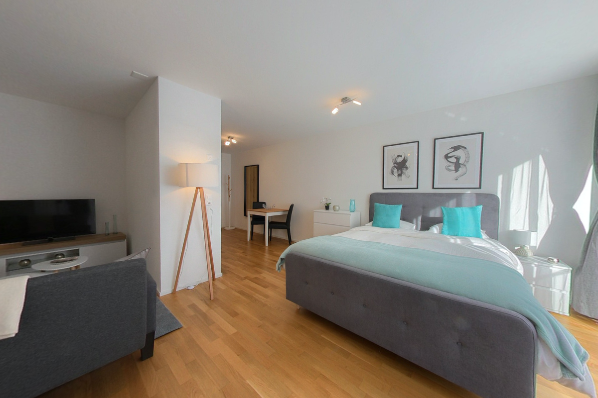 Large double bed with pillows, lamp, television, cabinet, small table, chair, hardwood floor.