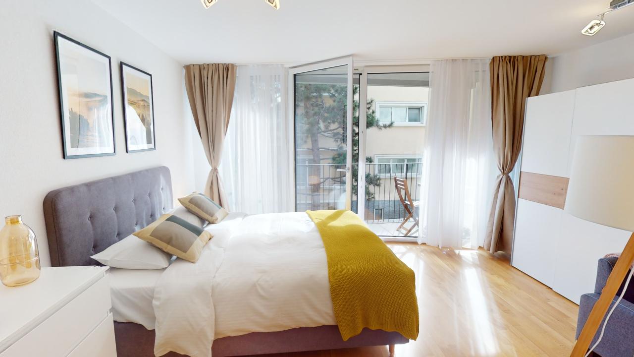 Spacious bedroom with large windows, balcony access, hardwood floors, gray upholstered bed frame, white bedding, yellow throw blanket, and framed artwork on the walls.
