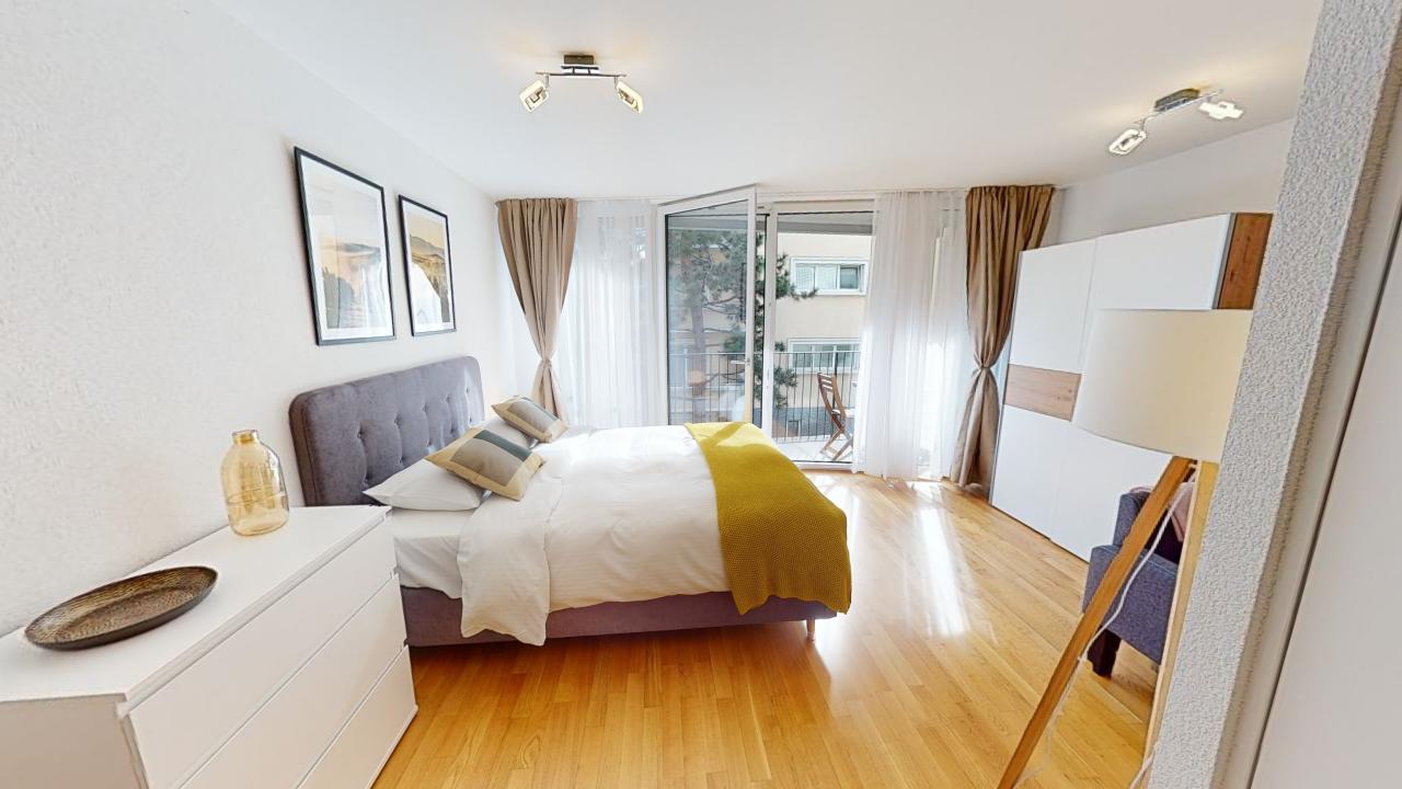 A spacious bedroom with hardwood floors, white walls, a bed, a nightstand, and an open balcony with curtains
