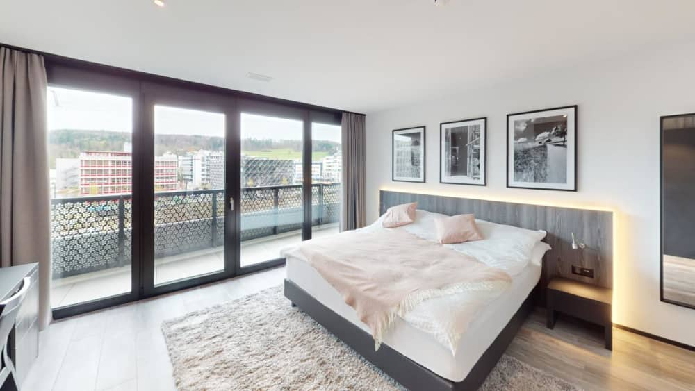 Large sliding glass doors, white bed, beige rug, bedside tables, mirrors, cityscape view