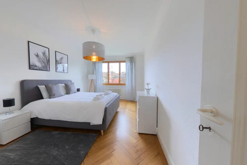Spacious bedroom with white walls, hardwood floors, large windows, gray bed frame, nightstands, and a decorative pendant light fixture
