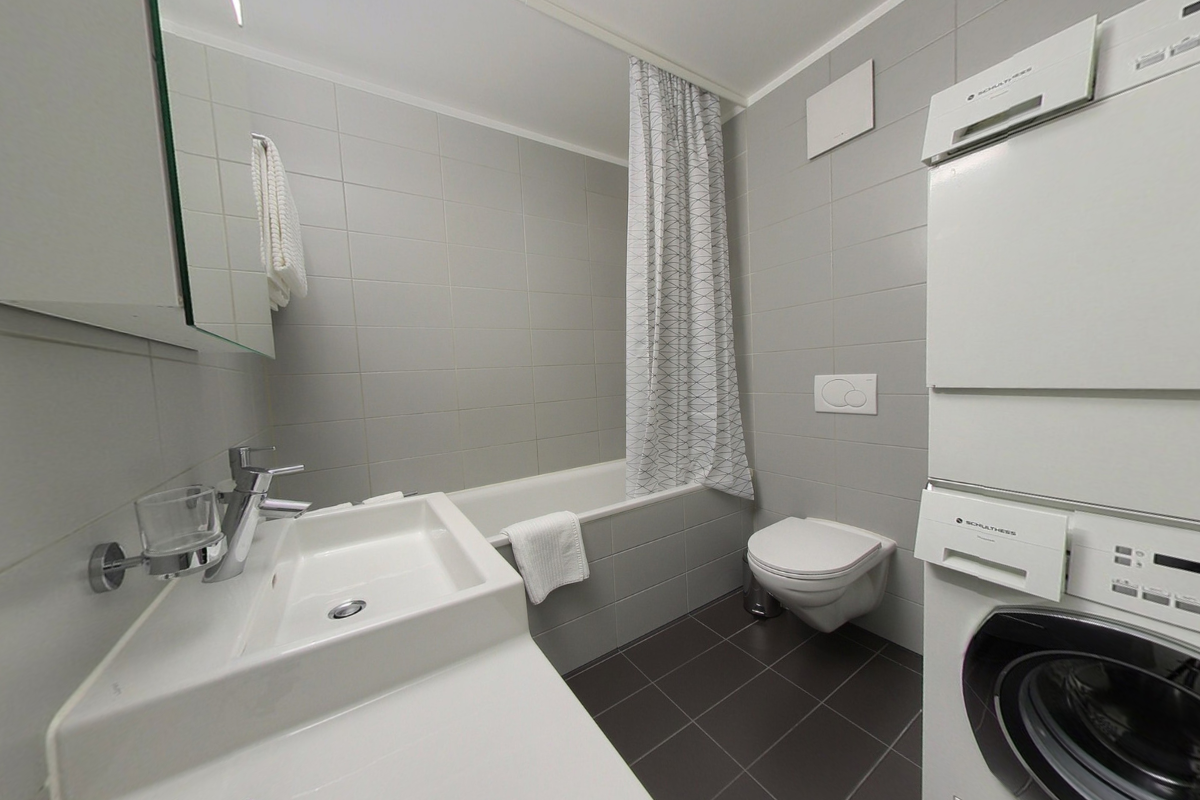 Spacious bathroom with gray tile walls and floors, a large sink vanity, a toilet, and a washing machine.