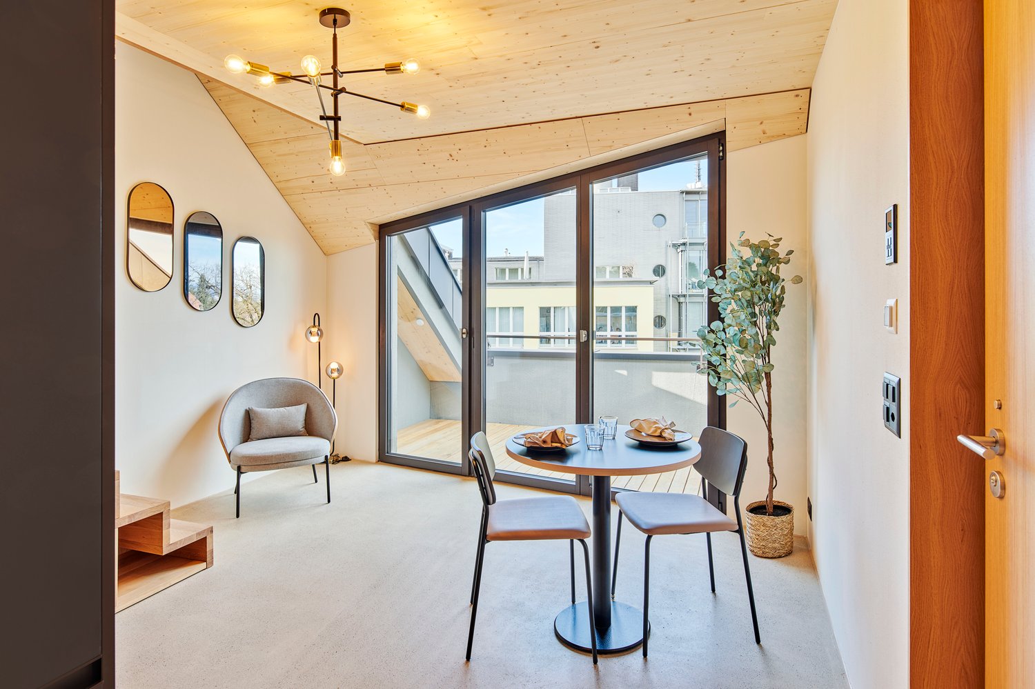 A room featuring round wooden ceiling, round mirrors on the wall, table and chairs with plants, glass door leading to balcony