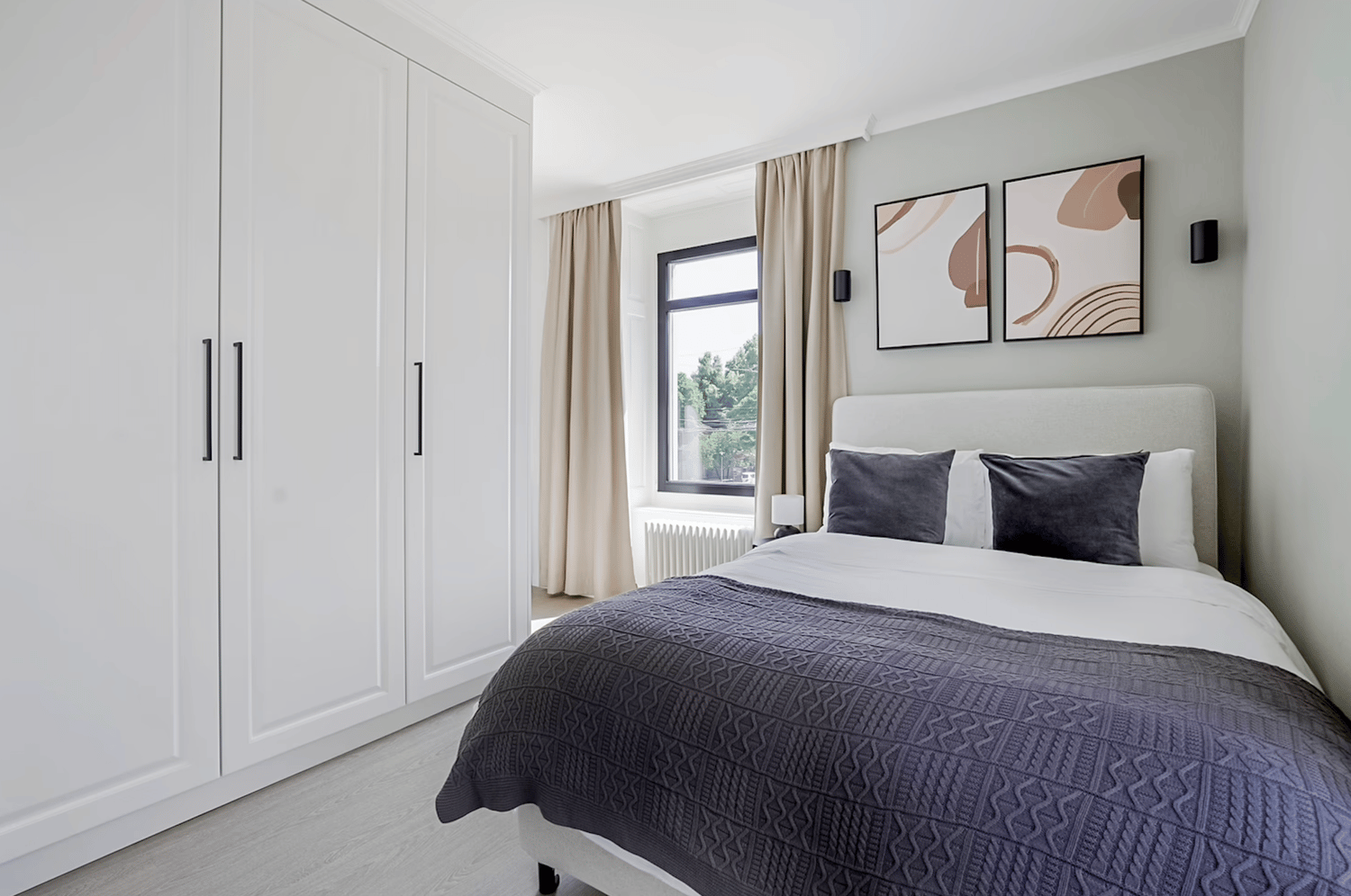 A contemporary bedroom with a white double bed and blue bedding, beige curtains, large white wardrobes, and two wall-mounted art pieces. A window with a view of trees and a white radiator complement the room's design.