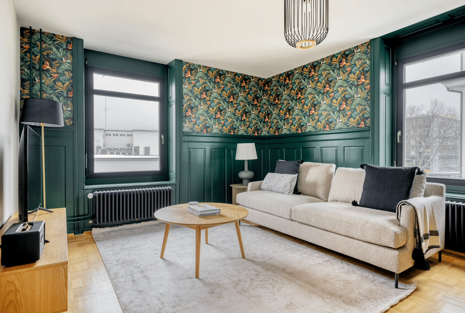 Spacious living room with large windows, green paneled walls, floral patterned wallpaper, beige sofa, wooden coffee table, and a pendant light fixture.