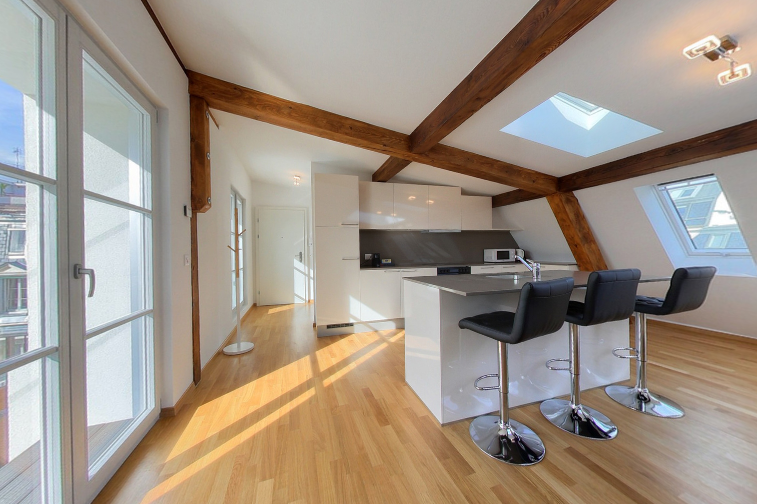 Open concept kitchen with white cabinets, black countertops, and wooden beams on the ceiling. The kitchen has a dishwasher and bar seating area with stools. The room has large windows allowing natural light to come in.