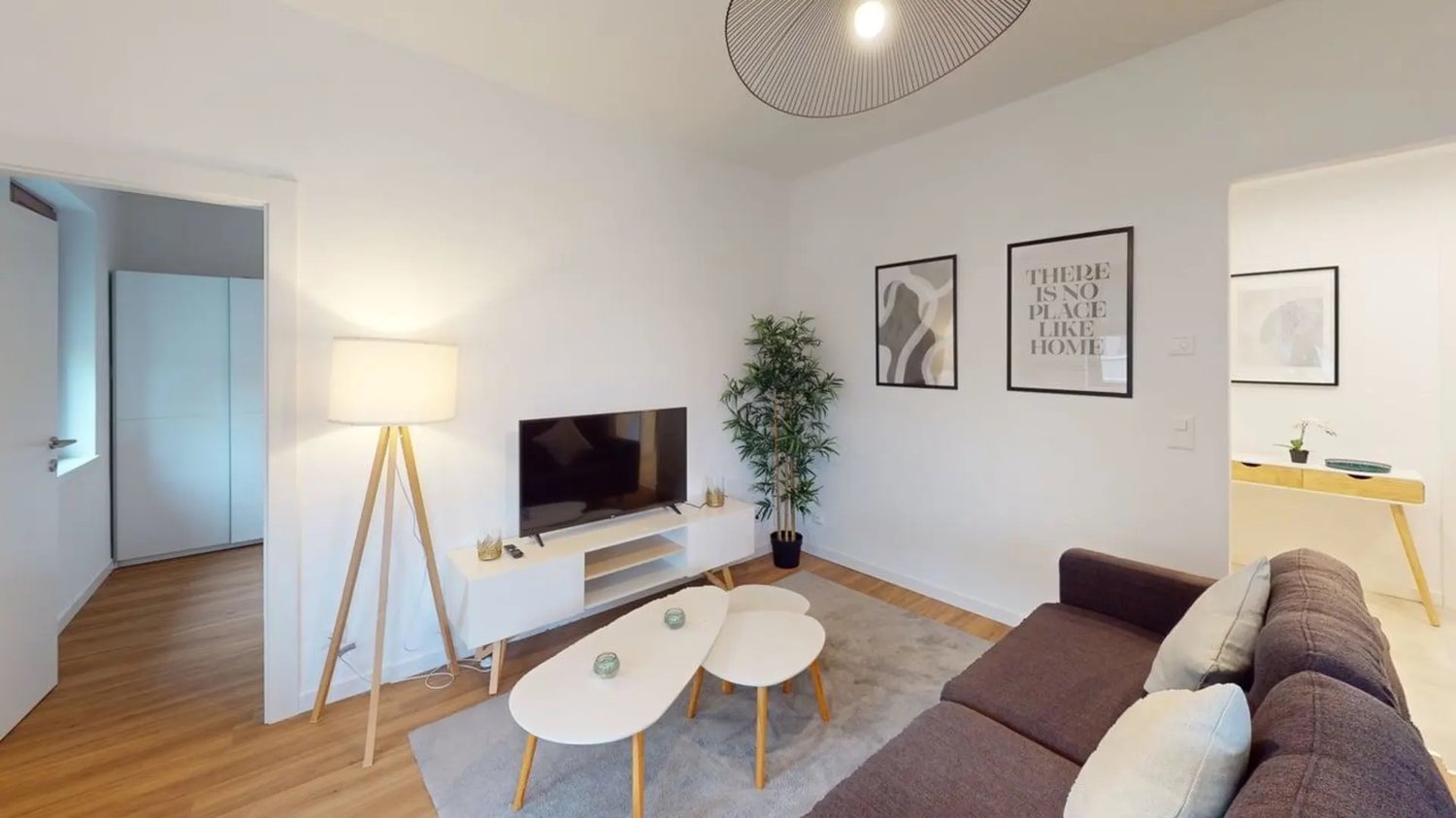 living room, white walls, wooden floor, brown sofa, two white tables, lamp, flat screen TV, wooden coffee table, carpet, plant, framed pictures