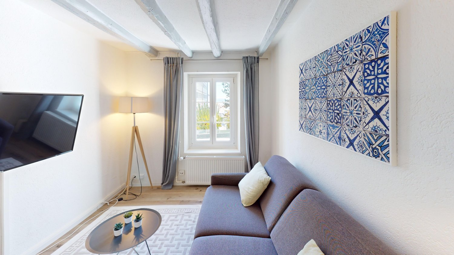 white walls, beige couch, round table, grey curtains, flat screen TV, lamp, tiled artwork, wooden floor