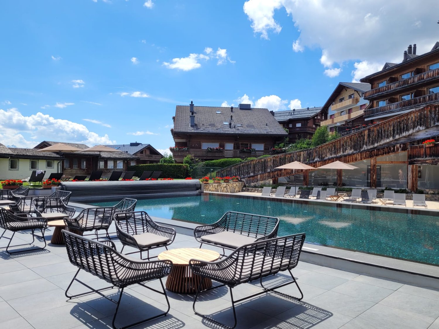 Modern pool area with lounge chairs, round wooden tables, umbrellas, and buildings in the background