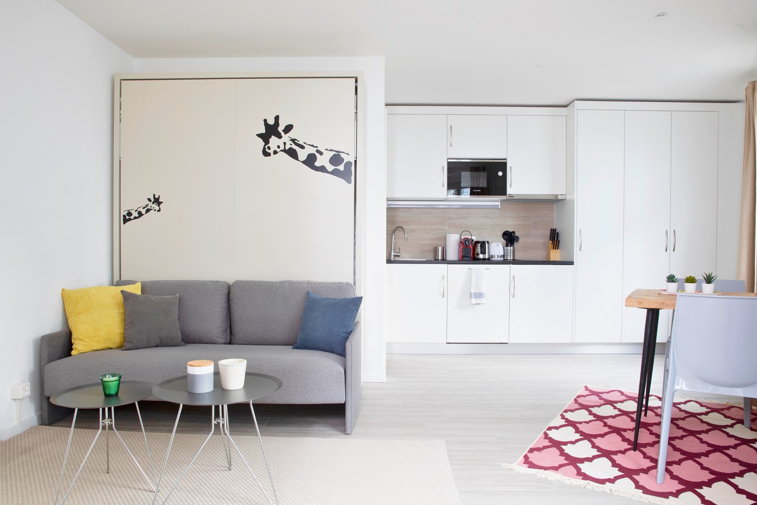 white living room with white walls, gray couch with pillows, white cabinets, wooden microwave, coffee machine, white coffee cups, carpet, white dining table with white chair, pink rug, white table with white cups