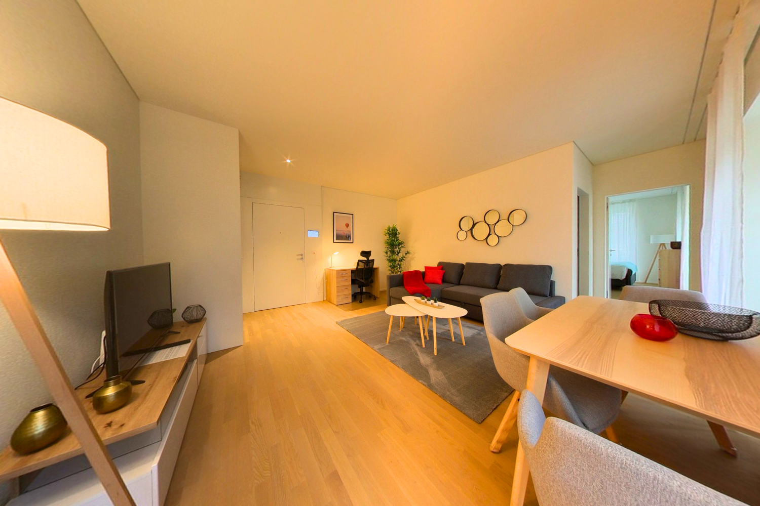 Spacious open-plan living room with hardwood floors, neutral-colored walls, large windows, modern furniture including a gray sofa, coffee table, and dining table, decorative wall art, and a TV stand.