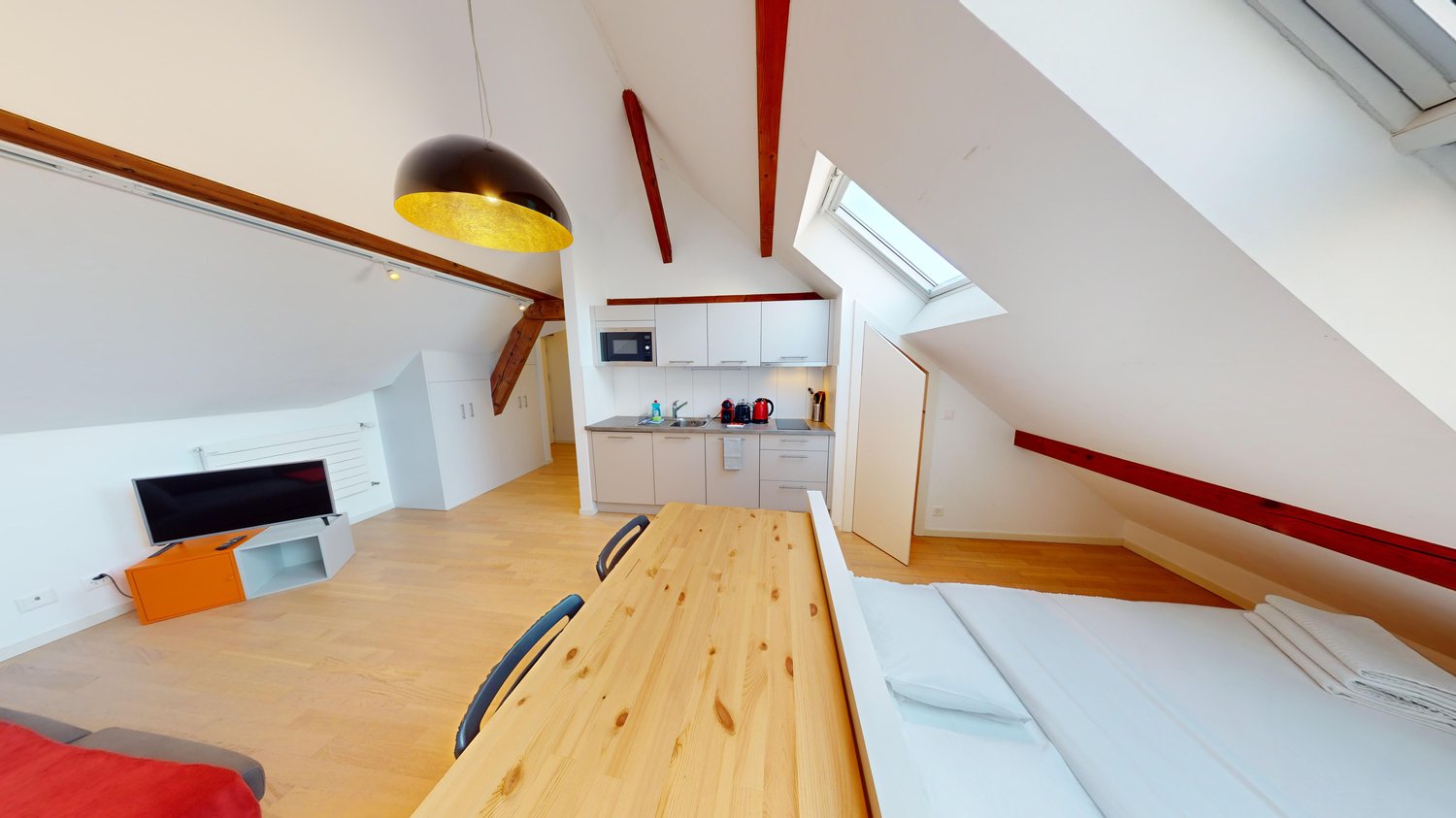 open floor plan, wooden table, two beds, one with a mattress, one with blankets, kitchen with sink and gas stove, microwave, refrigerator, and cabinets, large windows, attic beams, TV, hanging lamp