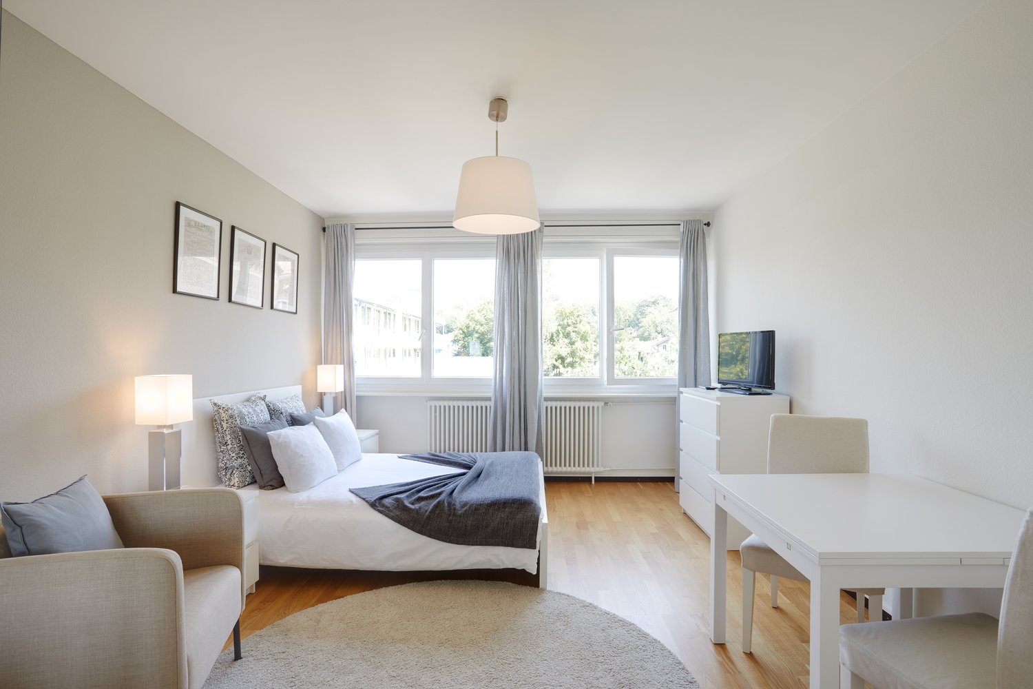 Bedroom with bed, pillows, blanket, lamps, TV, radiator, window, rug, chair, table, chairs