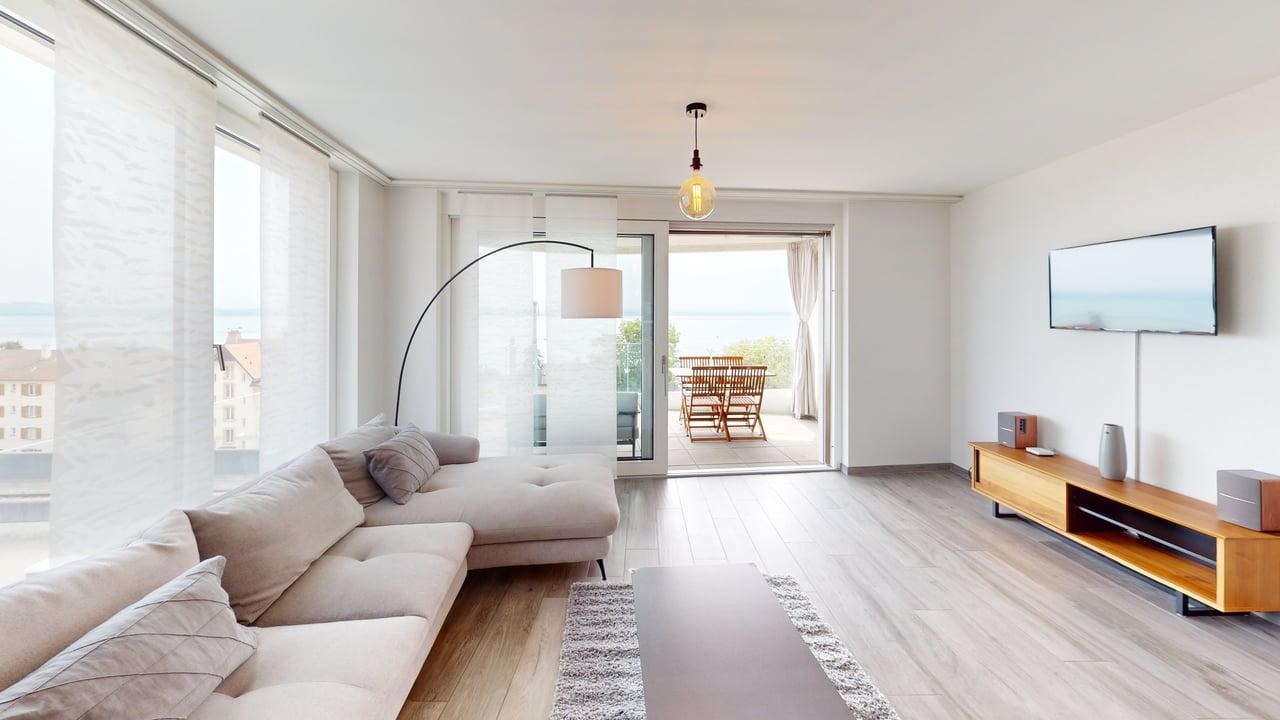 floor-to-ceiling windows, wooden floors, gray sofa, center table, floor lamp, tv stand, flat-screen TV, rug, large balcony with table and chairs