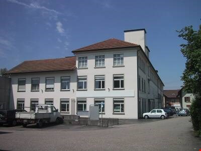 large white building, brown roof, several windows, white truck and car parked in front, other cars parked in the parking lot