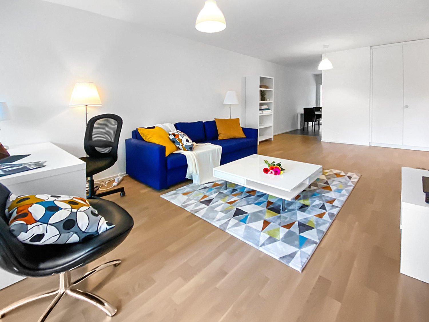 Spacious living room with hardwood floors, white walls, large windows, blue sofa, colorful patterned rug, white coffee table, and a black swivel chair