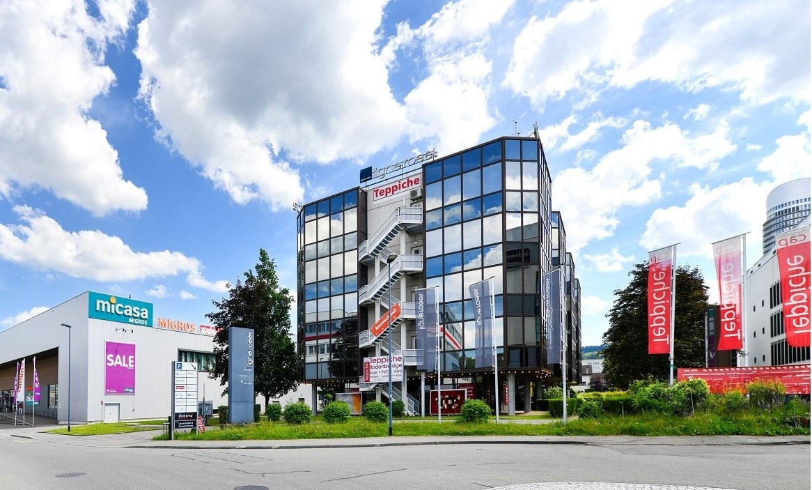 Multi-story commercial building with glass facade, Teppiche signage, and Micasa retail store. Outdoor parking area visible in front of the building. Flags and banners displayed on the building exterior.