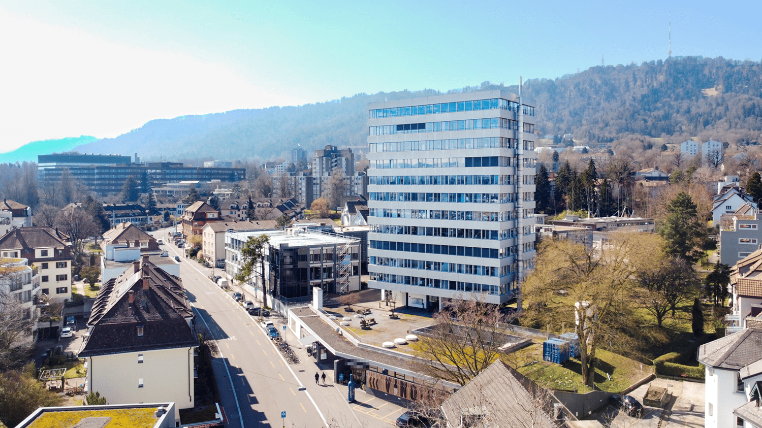 The image shows a cityscape with a mix of modern high-rise buildings and older, smaller structures. There are several multi-story office buildings with glass facades, as well as some residential buildings. The city is surrounded by hills and mountains in