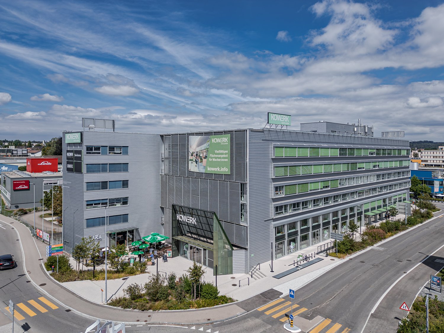 A large multi-story commercial building with the Kowerk logo and branding prominently displayed. The building has a modern, glass and steel design with several floors and a prominent entrance. There are also some smaller buildings and containers visible i