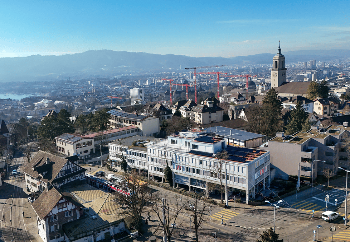Aerial view of urban buildings and infrastructure in a city, with buildings in various styles and sizes, surrounded by trees and a river, clear blue sky.
