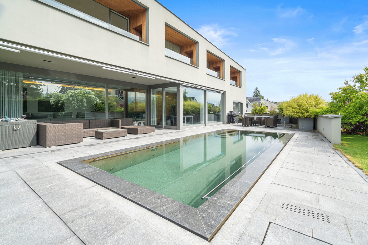rectangular swimming pool, grey tiled floor, sitting area with lounge chairs and tables, garden plants