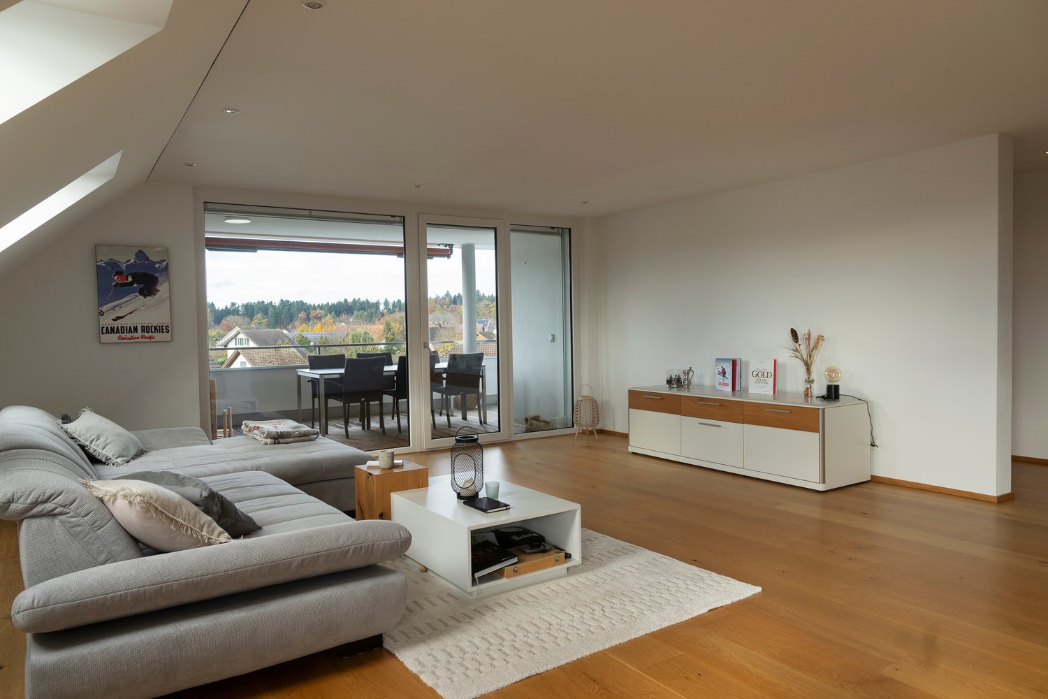 A modern and open living room with large windows and sliding glass doors leading to a balcony with an outdoor dining area and stunning views of the forest. The room features wooden floors, a comfortable grey sofa, a white coffee table, and decorative item