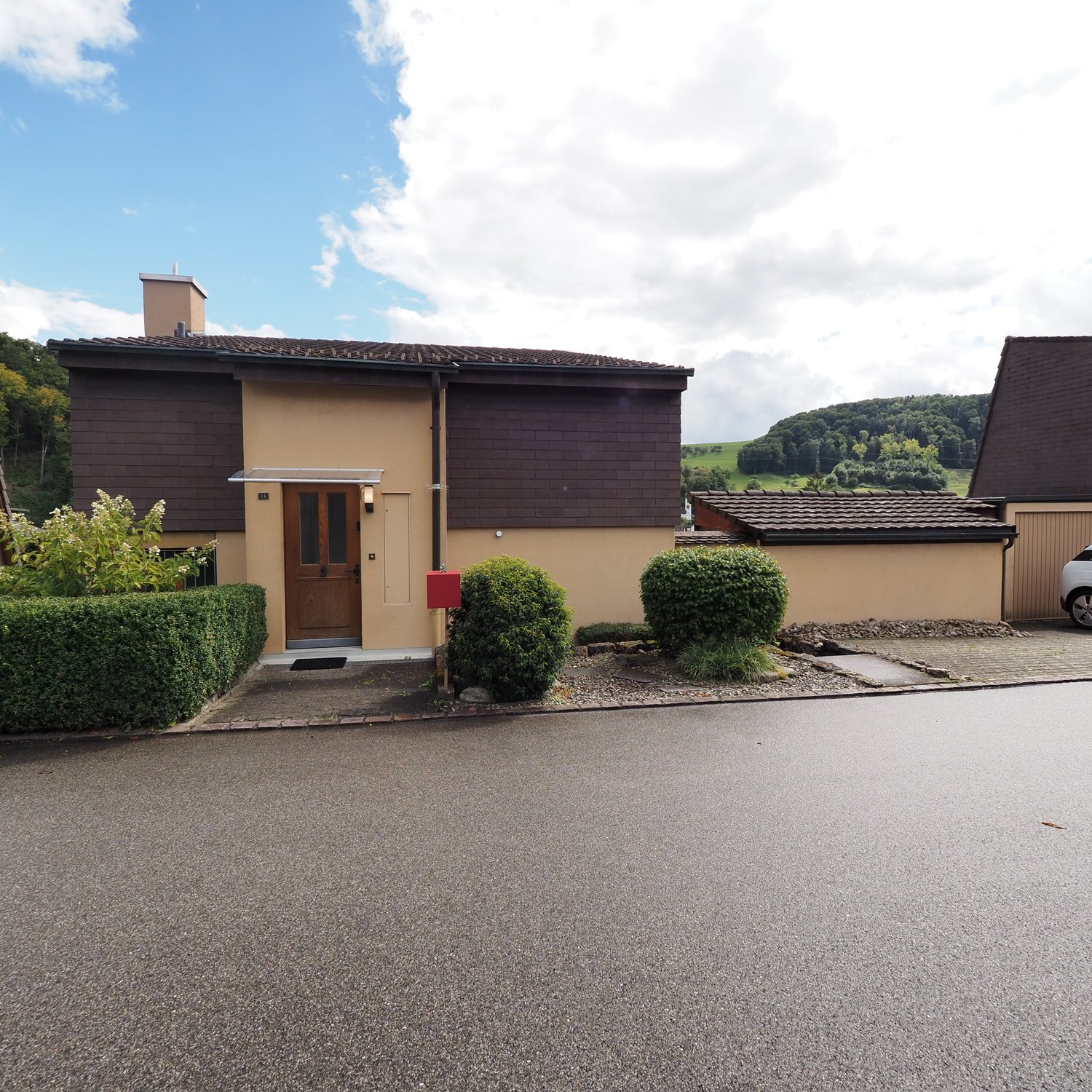 Brown and cream house, chimney, red mailbox, garage, gravel driveway, raised ground floor, greenery