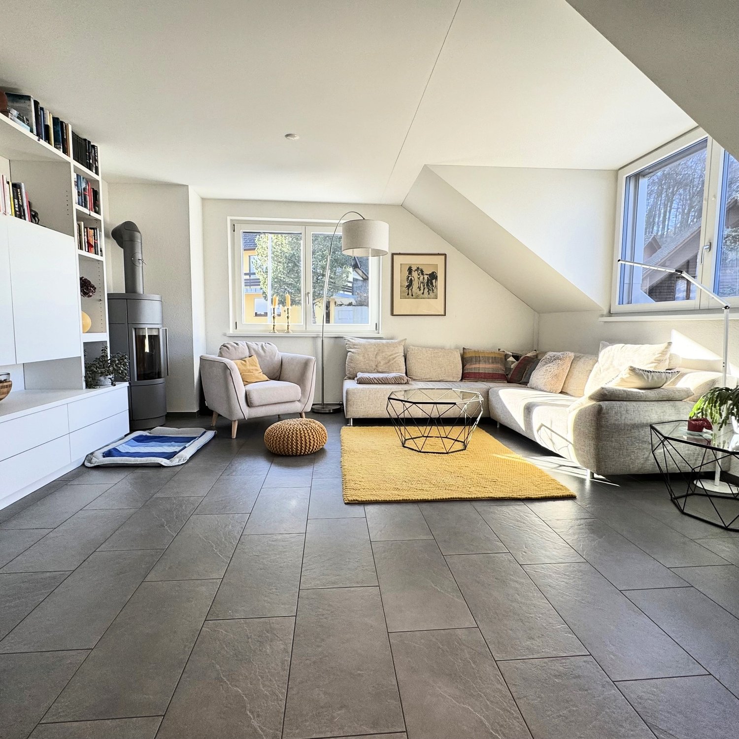 Gray flooring, beige sofa, two armchairs, center table, floor lamp, carpet, fireplace, shelf with books and plants
