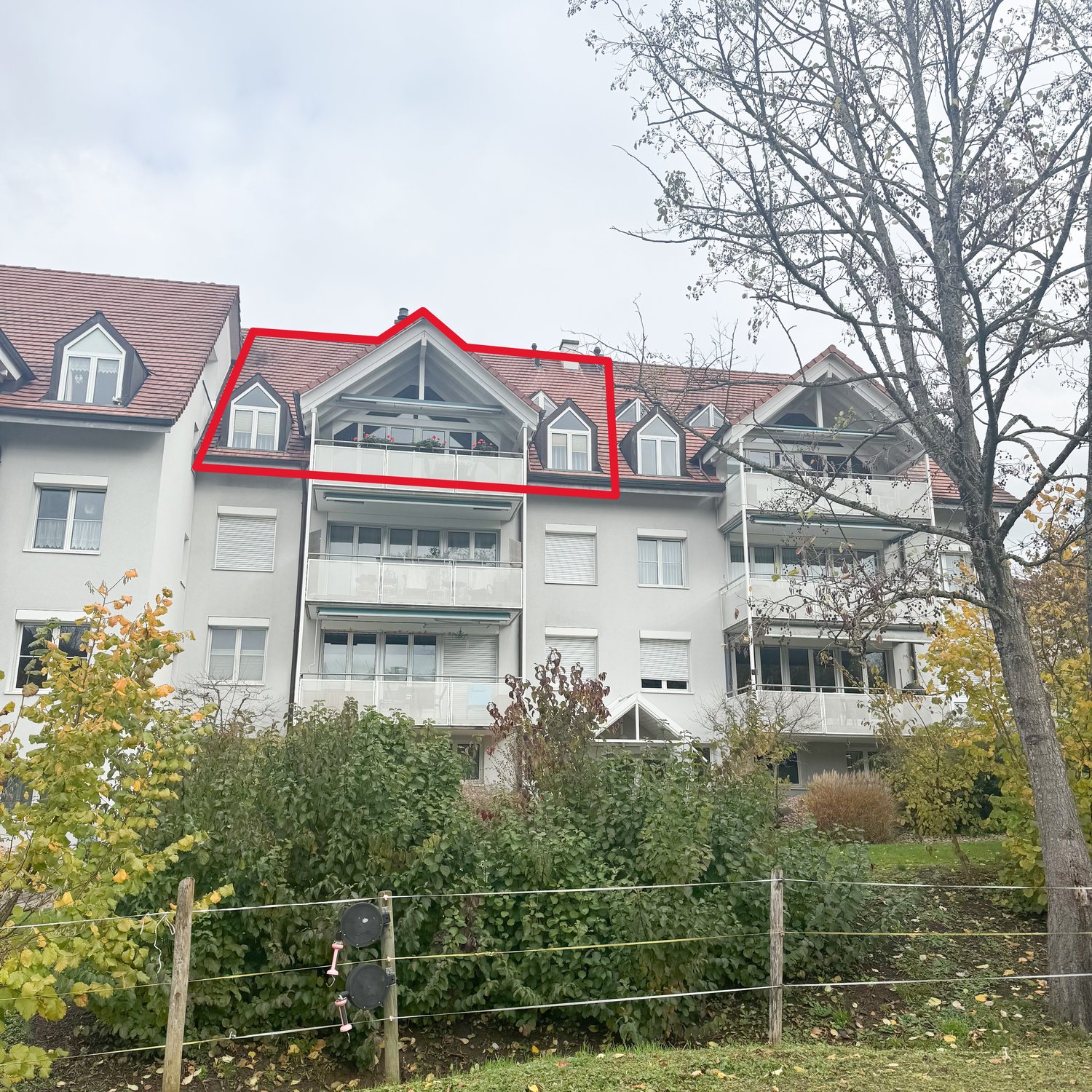 Apartment building with 4 stories, red roof, balconies, many windows, large front yard, greenery and plants, wooden fence, tree