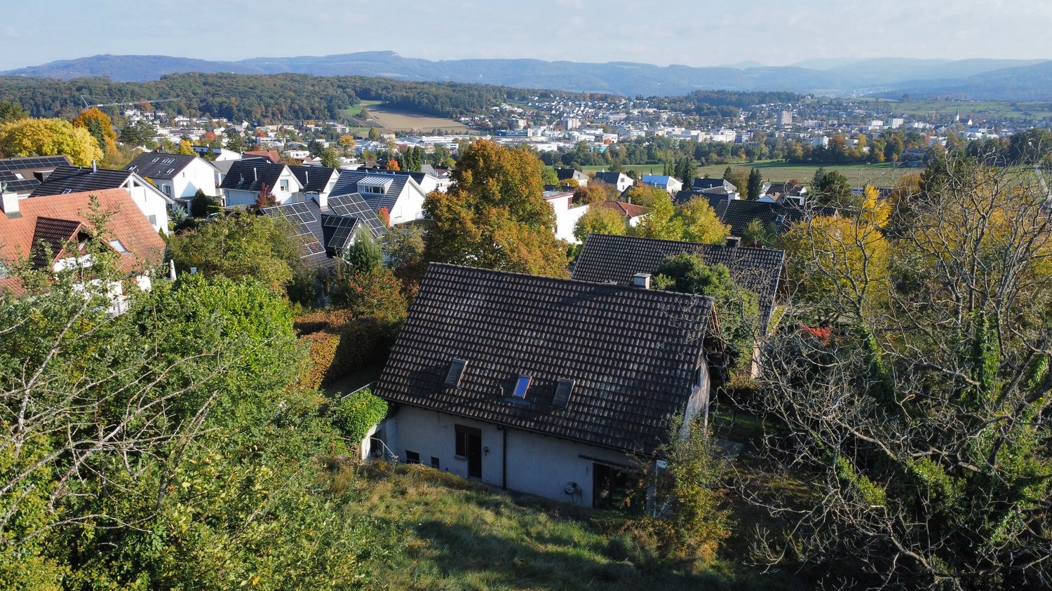 House in village, white walls, brown roof, surrounded by trees and shrubs, distant cityscape