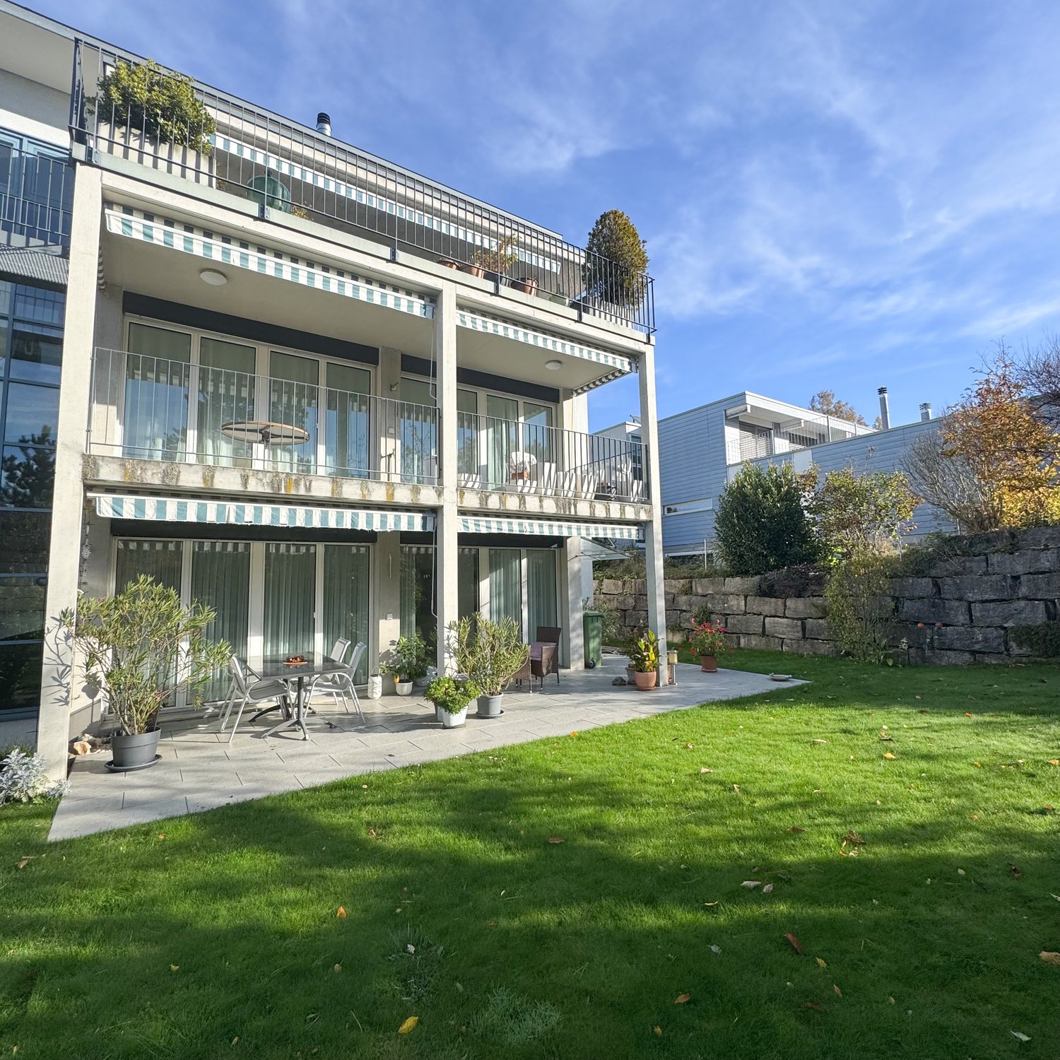 modern 2 story house, concrete walls, glass windows, balconies, outdoor table and chairs, green grass, garden, multiple potted plants