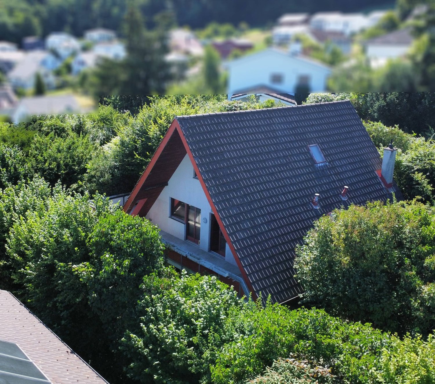 Two-story house with a red-tiled roof, surrounded by lush greenery and trees. The house has several windows and appears to have a balcony or terrace on the upper level.