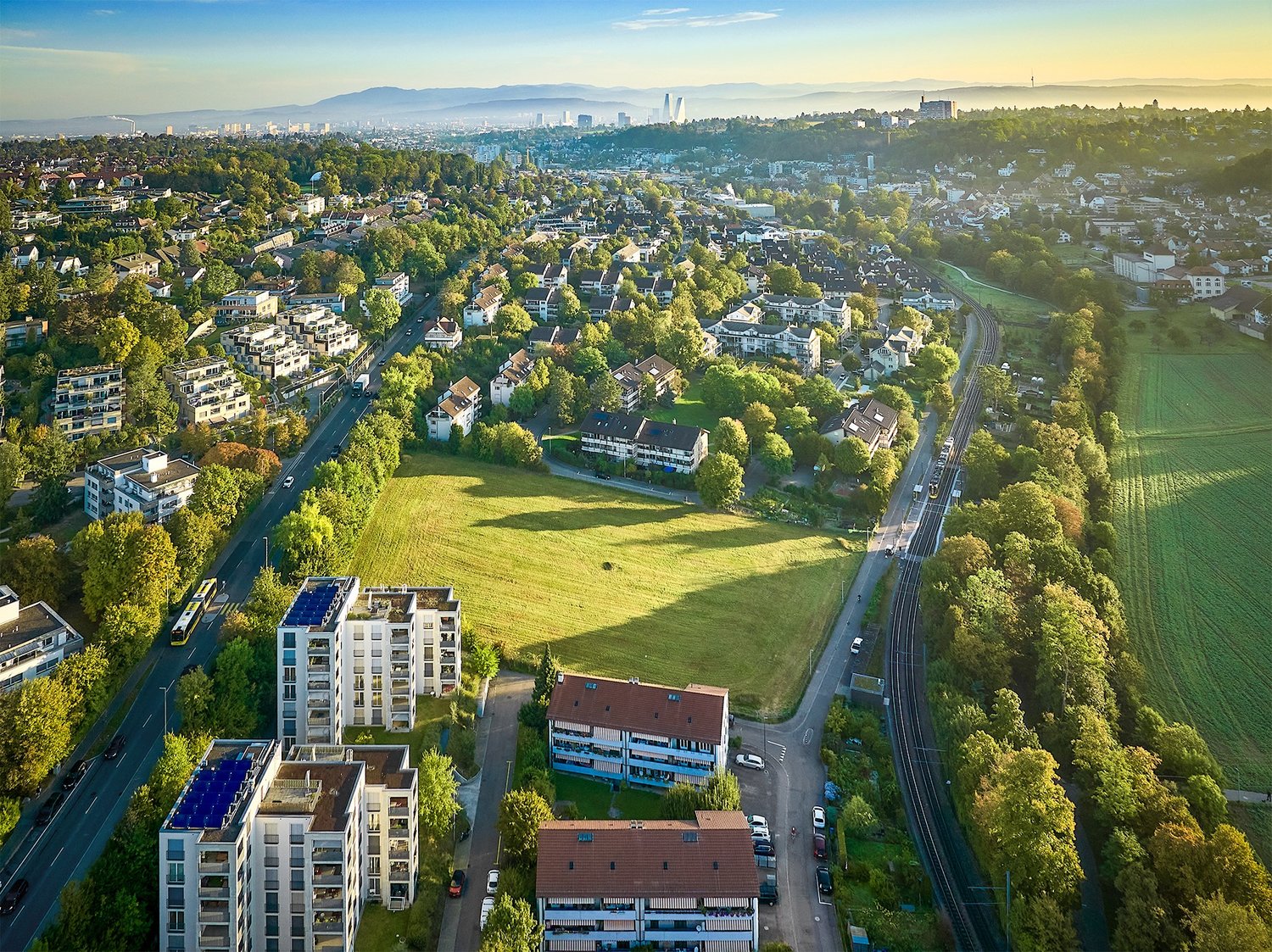 multiple buildings, roads, trees, and a large grassy field with a view of mountains