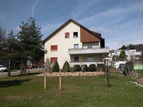 3-story apartment building with red and white exterior, balconies, and surrounded by trees and a grassy lawn