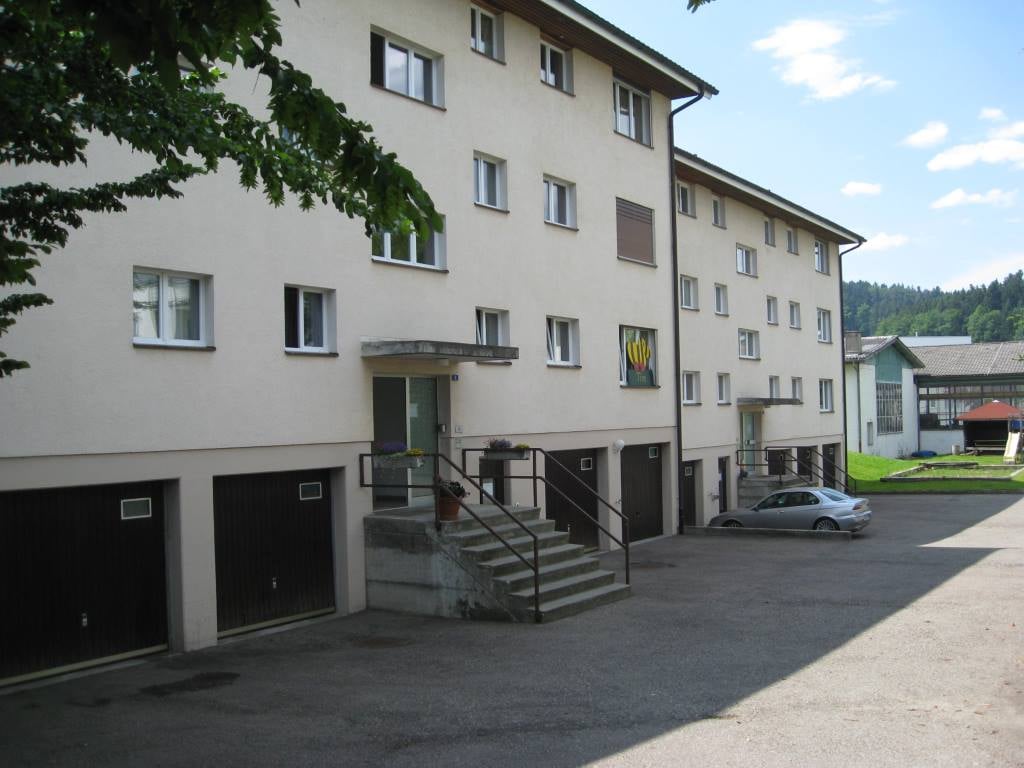 3-story apartment building with white exterior, multiple windows, and a garage and parking spaces in the front