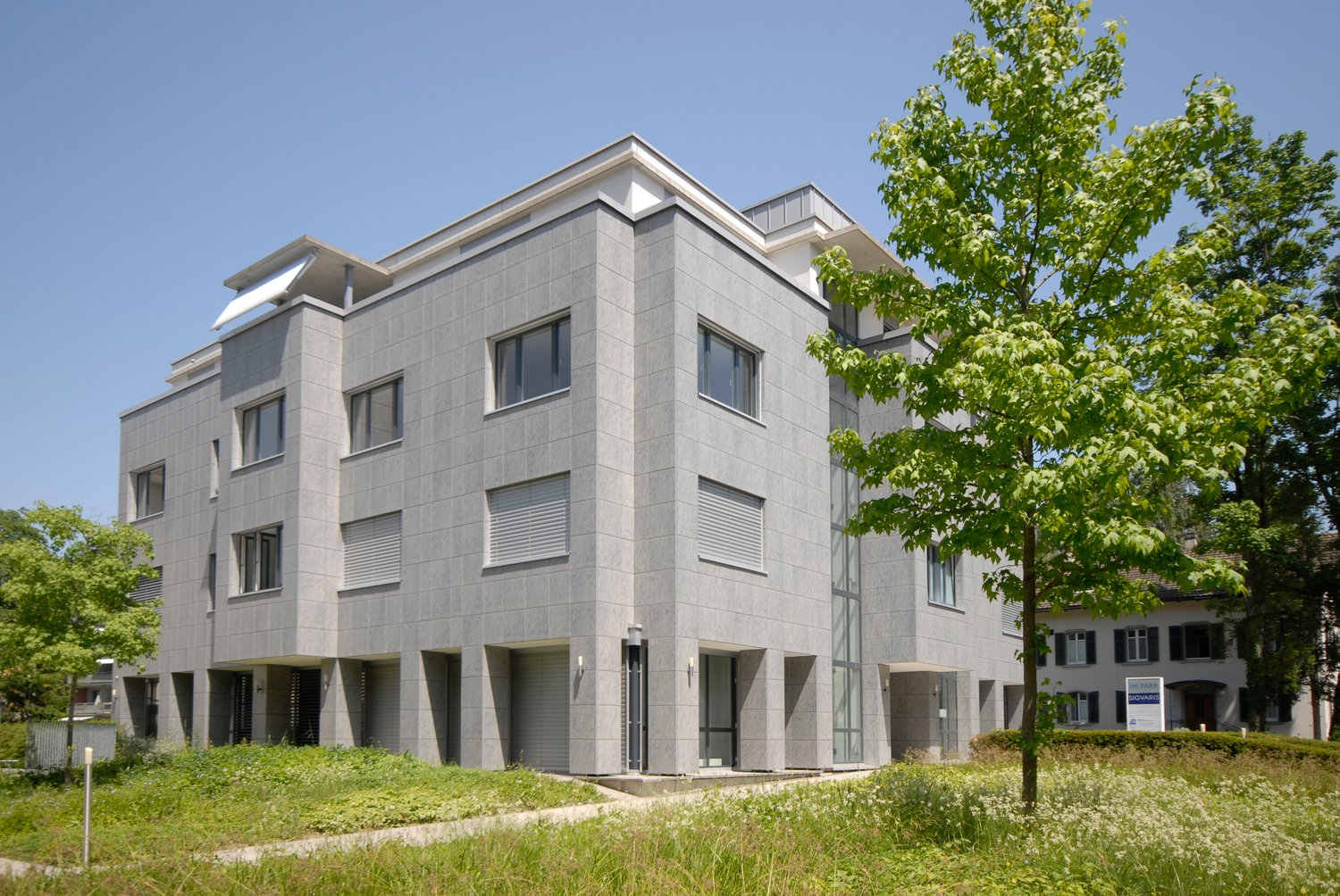 3-story modern office building with a gray facade, large windows, and a parking area in front. The building is surrounded by trees and greenery.
