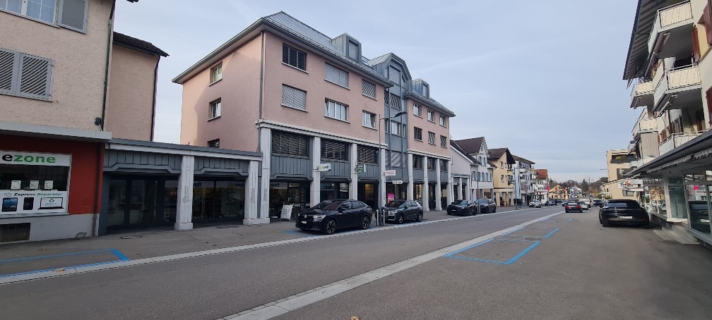 A street with a few parked cars in front of buildings, some with balconies and shops