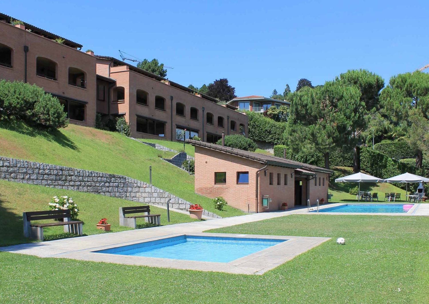 large brick building on the hill, large grassy yard, small pool, small house, benches, patio chairs and tables, large umbrellas